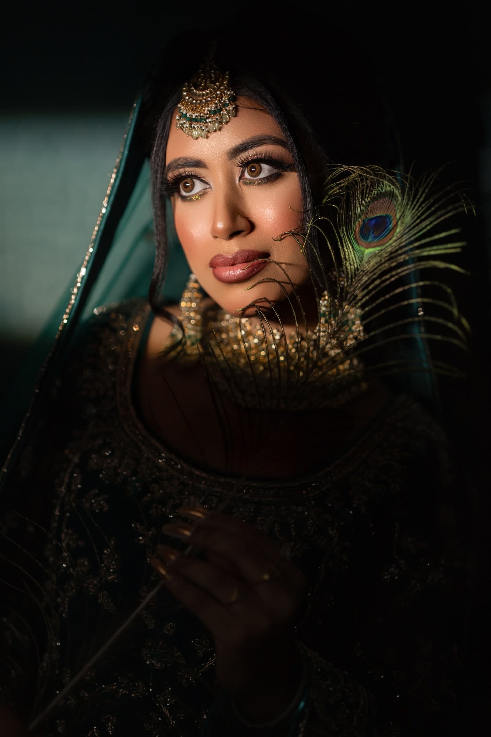 Artistic photoshoot portrait with moody lighting, emerald green veil, kundan jewellery and a peacock feather