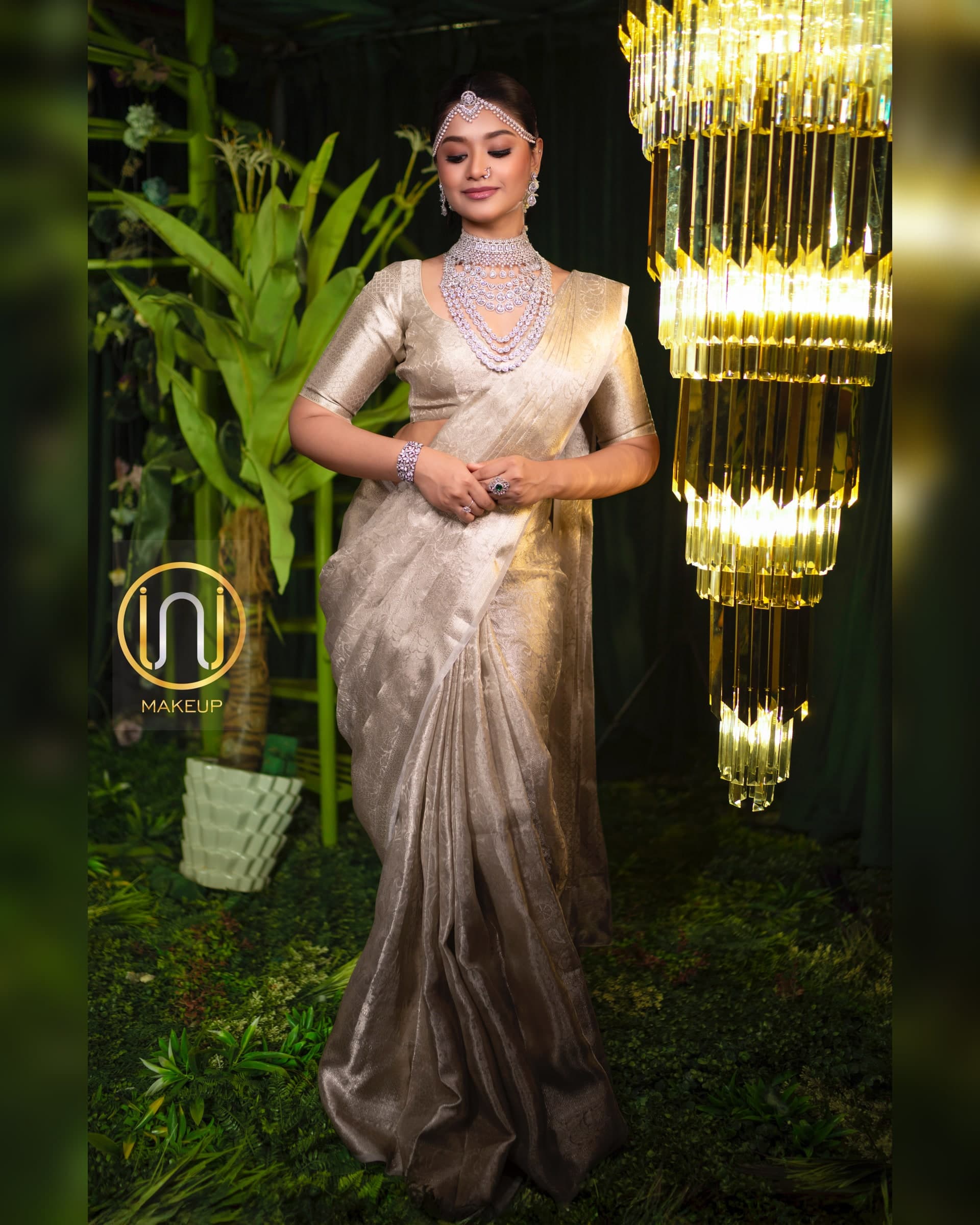 Full-length reception portrait in a metallic gold tissue saree with tiered diamond jewellery next to a chandelier
